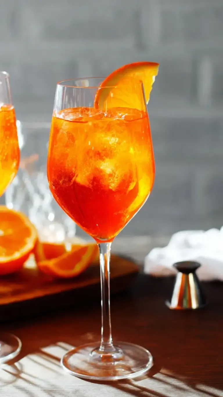 A vibrant orange Aperol Spritz cocktail in a large wine glass, garnished with an orange slice, on a sunny patio table.