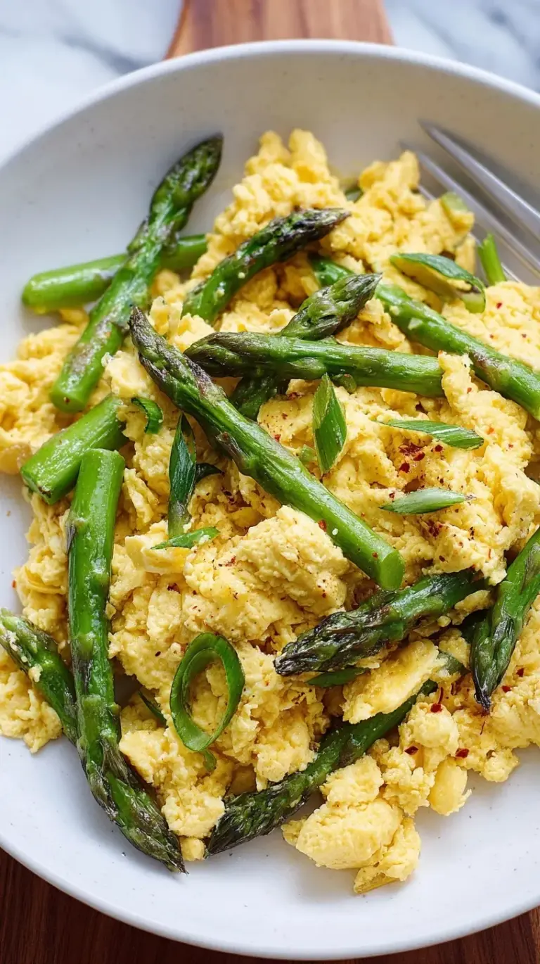 A close up of soft-scrambled eggs with bright green asparagus pieces and fresh chives on a white plate.